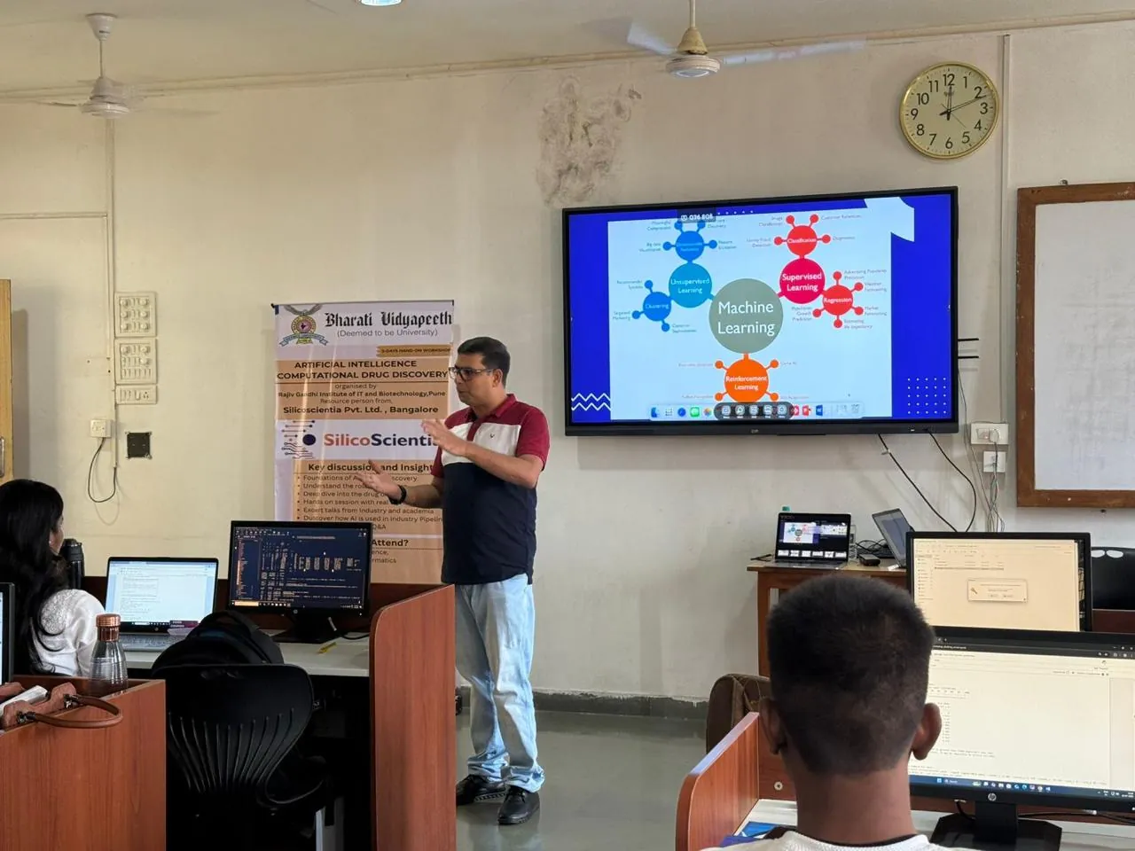 An instructor explaining machine learning concepts to a computer laboratory, with a workshop banner and a slide on screen