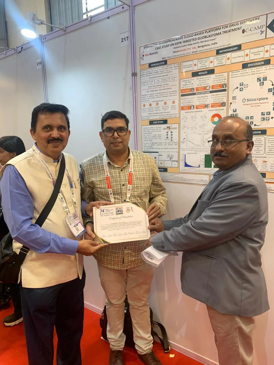 Three men holding a certificate of presentation in front of a research poster at the 74th Indian Pharmaceutical Congress