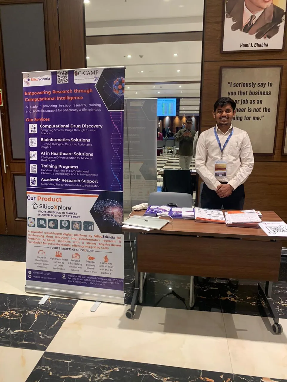 A team member at the SilicoScientia conference stand beside a banner listing training and academic research support